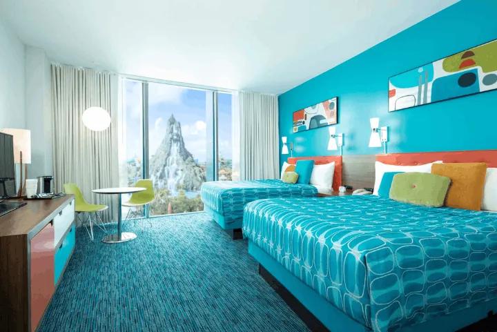 A retro-themed hotel room with two queen beds, bright teal walls, and patterned blue bedspreads, featuring a large floor-to-ceiling window that offers a direct view of the Volcano Bay water park volcano