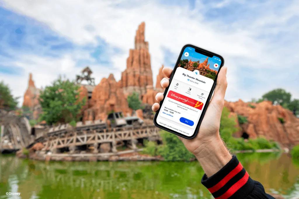 A hand holding a phone showing the Disney Premier Access booking screen for Big Thunder Mountain, with the ride's rock formations in the background