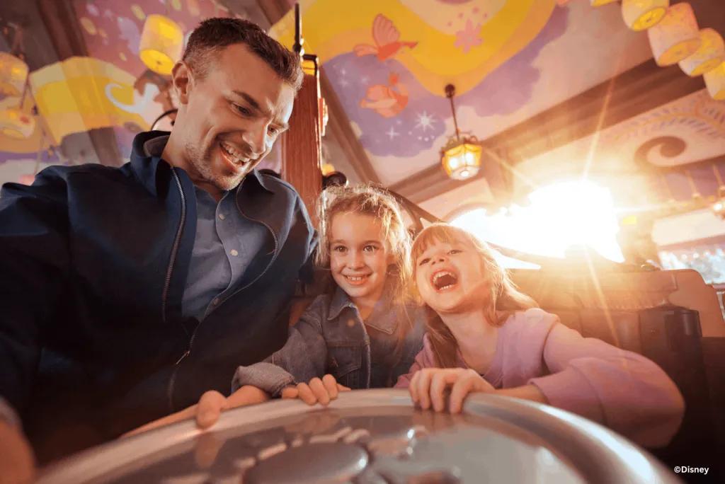 A father and two laughing daughters spin together on Raiponce Tangled Spin, lit by golden sunlight beneath a lantern-decorated ceiling at Disney Adventure World