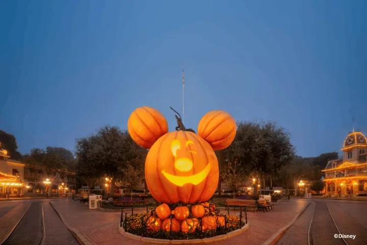 A giant glowing Mickey Mouse jack-o'-lantern display centered on Main Street, U.S.A. at Disneyland Resort during dusk with illuminated Victorian buildings in the background