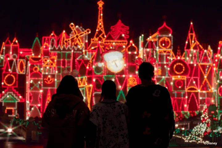 Silhouettes of three guests wearing Mickey Mouse ears looking at the illuminated "it's a small world" Holiday façade covered in festive red, green, and gold lights at night