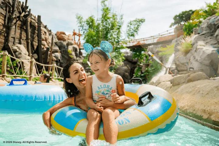 A mother and young daughter wearing sparkly blue Mickey ears smile together while riding an inner tube down Castaway Creek at Disney's Typhoon Lagoon Water Park