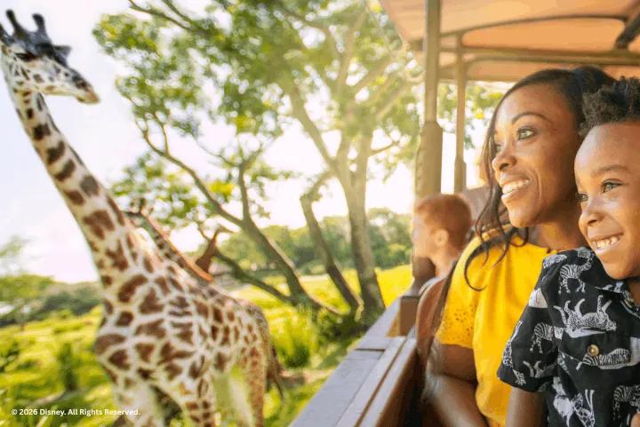 A mother and child smiling at a giraffe up close from a safari vehicle on Kilimanjaro Safaris
