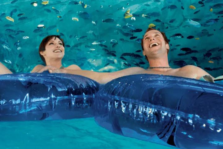 A smiling man and woman floating on dark blue inner tubes through an underwater clear acrylic tunnel surrounded by tropical fish and marine life