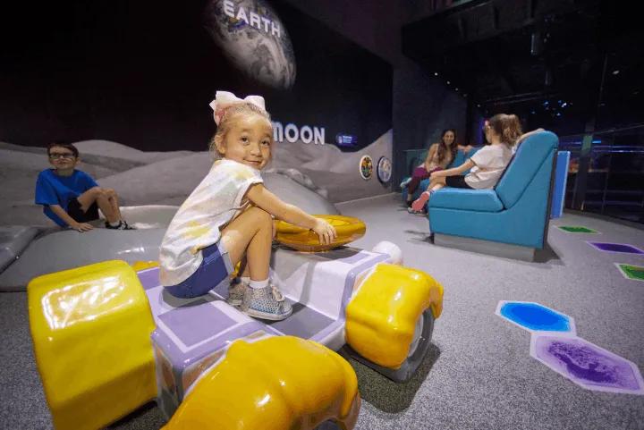 A young girl riding a toy rover inside the Planet Play area