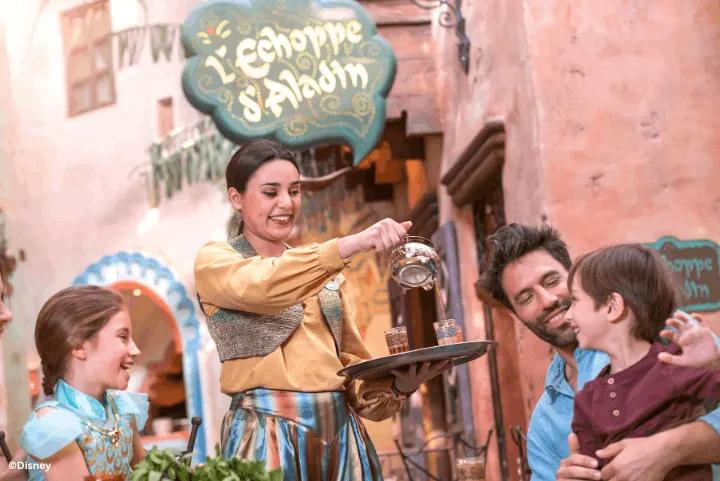 A Disneyland Paris cast member pouring mint tea into glasses on a tray for a smiling family seated outside L'Échoppe d'Aladin in Adventureland