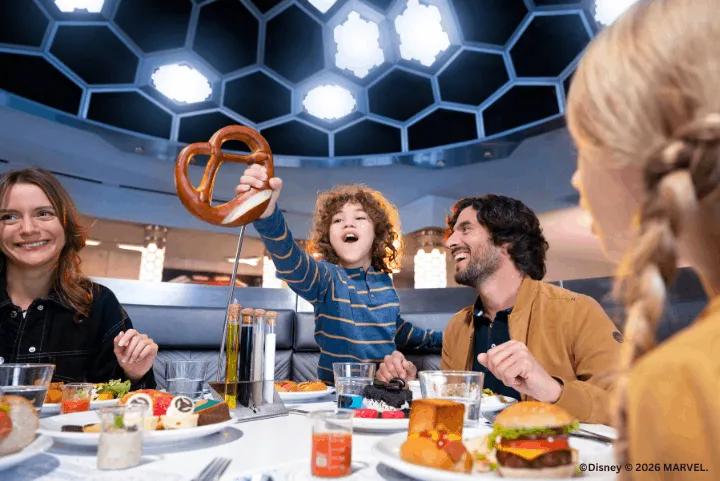 Family laughing at a table as a child holds up a large pretzel at PYM Kitchen, Avengers Campus