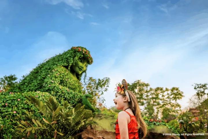 A young girl dressed in a Moana costume smiles up at a large Te Fiti topiary covered in green foliage and pink flowers