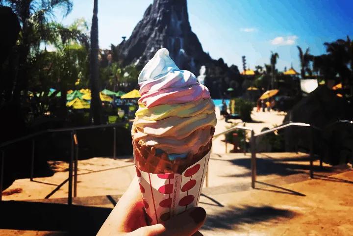 Soft-serve ice cream cone swirled with colorful pastel layers, held against the backdrop of Volcano Bay's Krakatau volcano