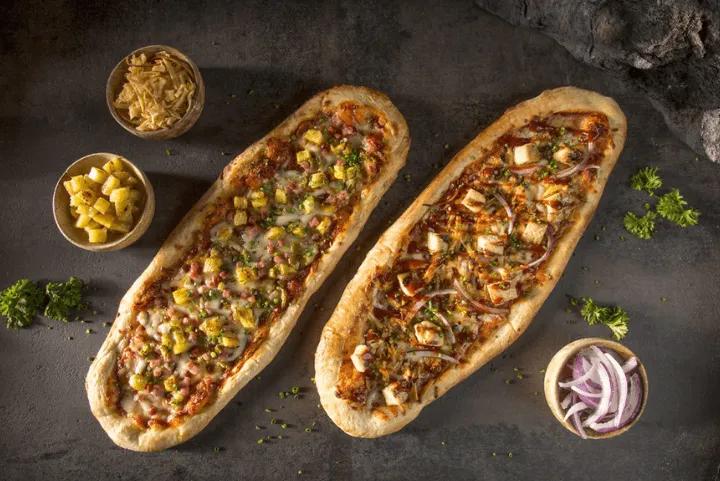 Two surfboard-shaped longboard pizzas with Hawaiian and BBQ chicken toppings next to small side bowls