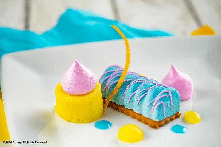 Plated colorful dessert with a blue log-shaped cake, pink meringue kiss, and yellow cake cylinder