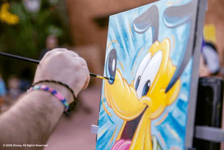 Hand using a fine paintbrush to detail a colorful canvas painting of Pluto