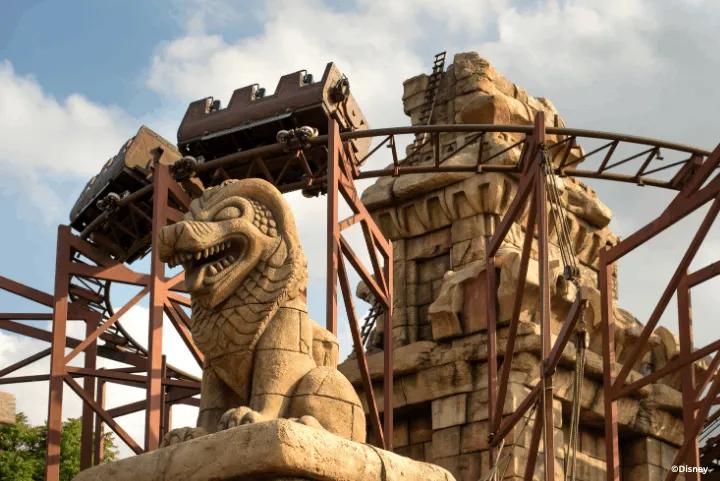 A mine train roller coaster looping through ancient stone ruins past a large stone lion statue