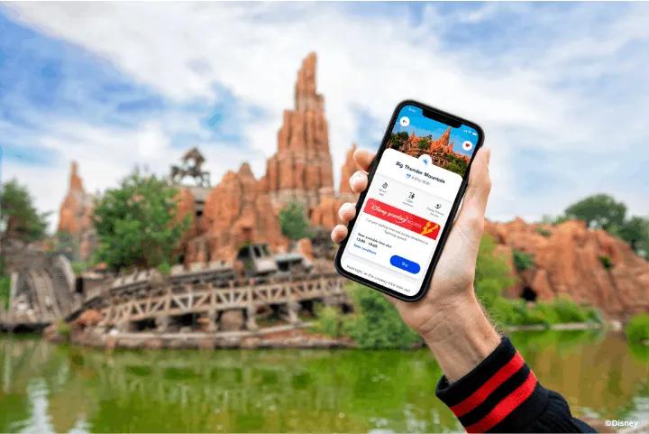 A hand holding a smartphone showing the Disney Premier Access feature in front of Big Thunder Mountain