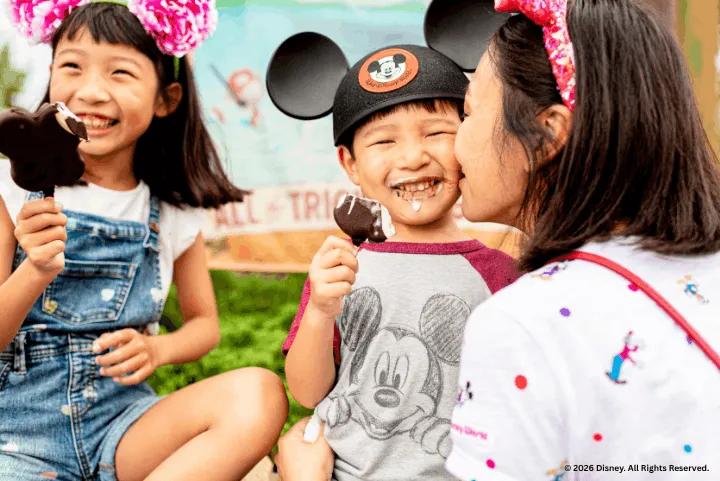 A mother and two children wearing ear hats laughing while eating Mickey Mouse ice cream bars