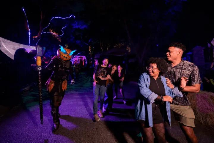 Guests reacting to a pumpkin-headed scareactor holding an axe in a outdoor scare zone at night