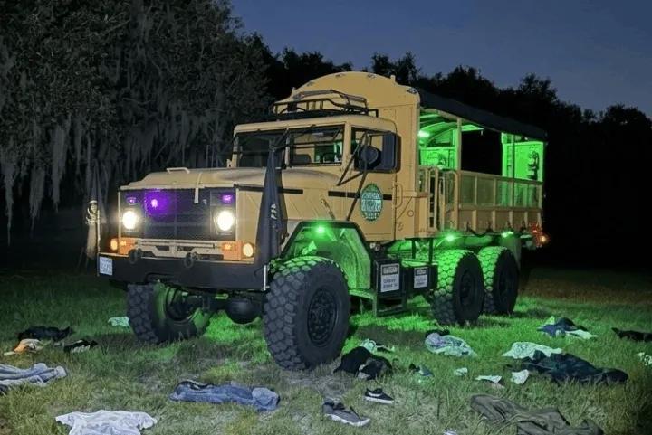 A tan military transport truck with green underglow lighting stands in a dark field covered with scattered clothes at the Terror 2 event