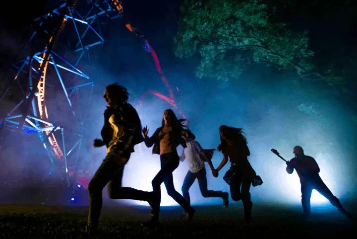 Silhouetted guests running away from a scareactor near a illuminated roller coaster at night