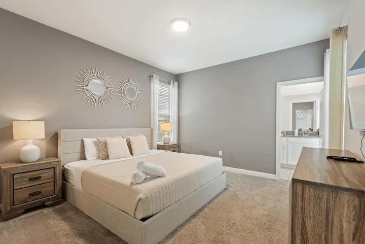 A gray-walled bedroom with a beige upholstered bed, wooden nightstands, sunburst wall mirrors, and an open doorway to an ensuite bathroom