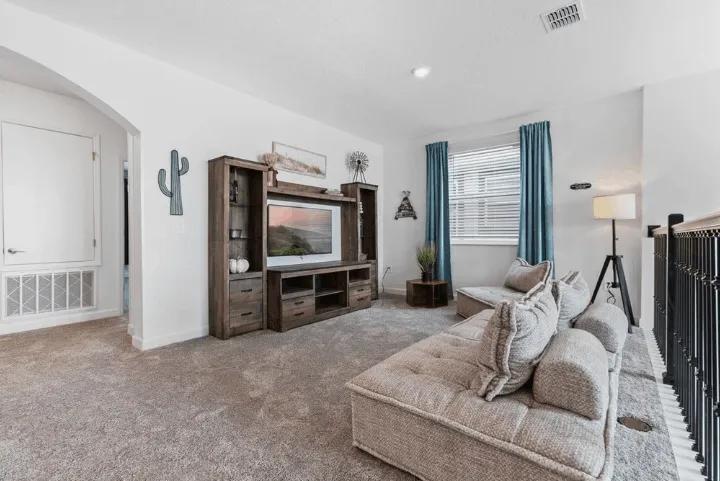 An upstairs loft living area featuring carpeted flooring, a low-profile sectional sofa, dark wood entertainment center, and blue accent curtains