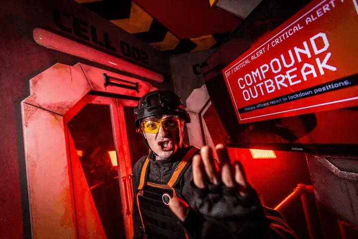 A Phalanx guard in tactical gear gesturing stop under a red emergency light near a screen reading COMPOUND OUTBREAK