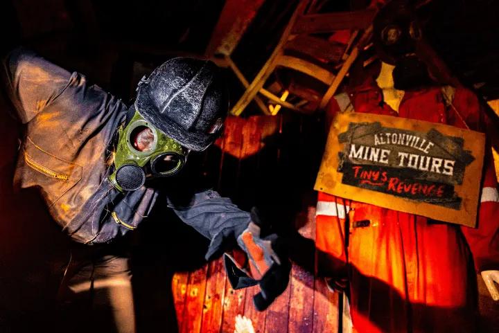 A actor wearing a gas mask and hard hat reaching out inside a dark mine maze next to a sign for Altonville Mine Tours: Tiny's Revenge