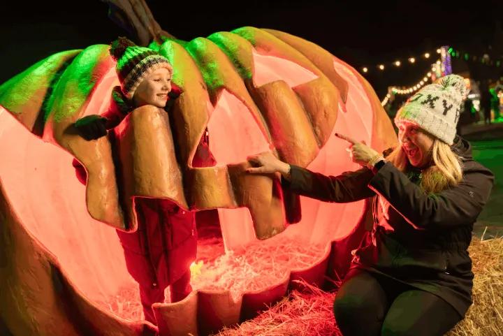 A child standing inside a giant illuminated pumpkin sculpture while a woman points and laughs