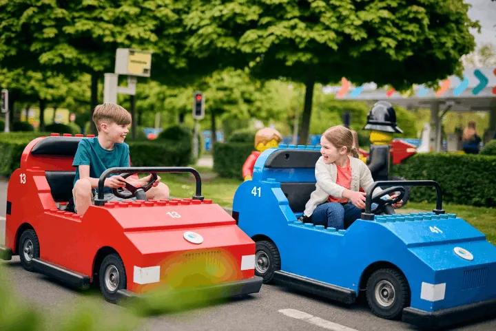 Two children smiling while driving red and blue LEGO cars