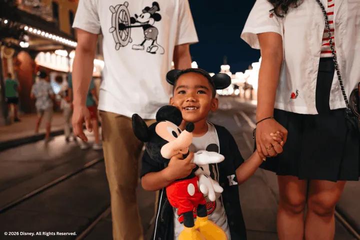 A young boy wearing Mickey Mouse ears holds a Mickey plush while walking with his parents along Main Street, U.S.A. at Magic Kingdom Park at night