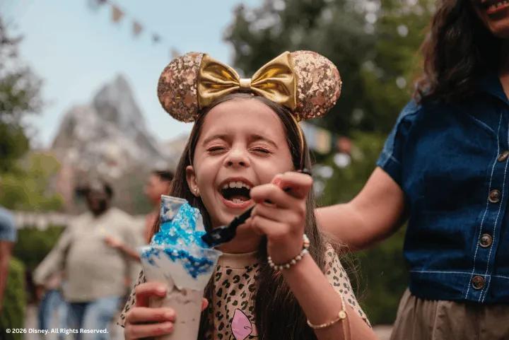 A young girl wearing sparkly Minnie Mouse ears laughs happily while eating a blue Yeti Sundae at Disney's Animal Kingdom Theme Park