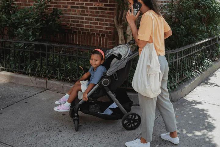 A mother talking on her phone while standing beside her young child sitting in a stroller on a sidewalk