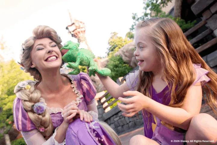 A young girl holding a green Pascal plush toy playfully touches Rapunzel's cheek outdoors at a Disney theme park
