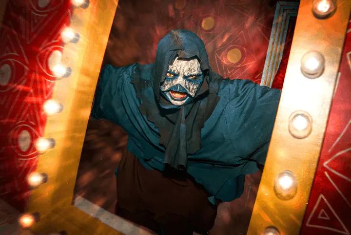 A hooded, masked figure leaning through a lightbulb-lined frame