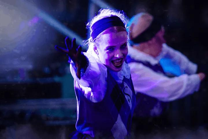 A performer in a school uniform with dark makeup reaching toward the camera under spooky blue lighting