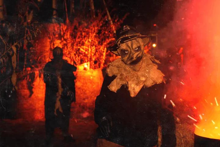 A person wearing a creepy scarecrow mask and a black hat, standing in a dark outdoor area illuminated by bright red light and sparks
