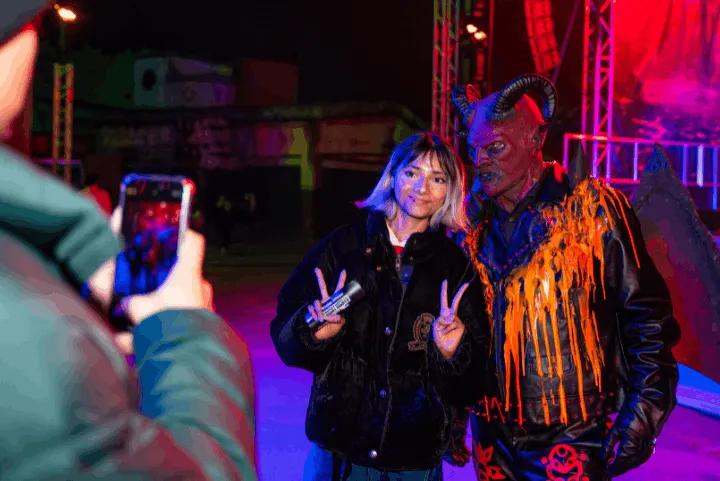 A guest posing for a smartphone photo with a horned scare actor at a brightly lit night event