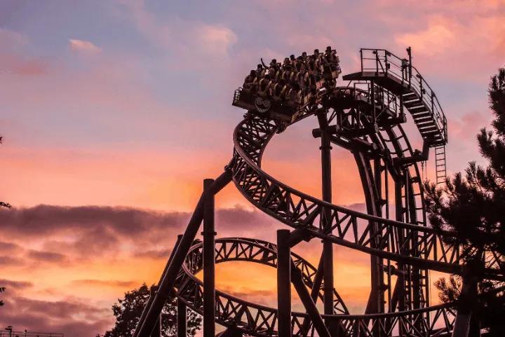 A rollercoaster car full of riders on a looping track silhouetted against a vibrant orange and pink sunset