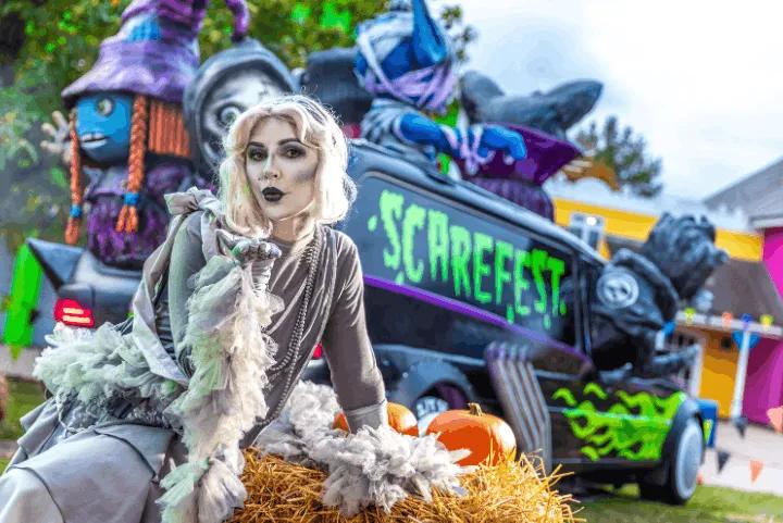 A costumed actor sitting on a hay bale blowing a kiss in front of a decorated Scarefest vehicle