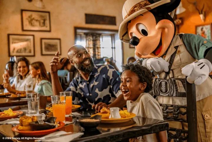 Mickey Mouse in safari gear interacting with a smiling young boy and his family at a restaurant table