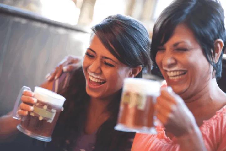 Two women laughing with foam moustaches while holding mugs of Butterbeer at The Leaky Cauldron