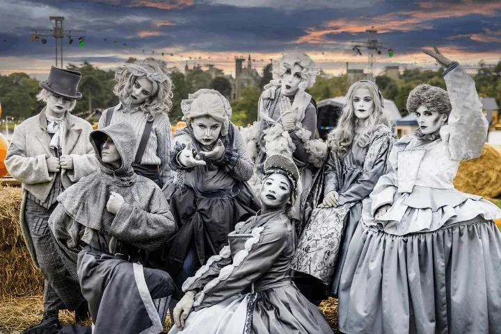 A group of actors in vintage costumes and pale makeup posing together outdoors