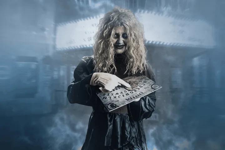 A spooky scare actor holding and pointing at a Ouija board in a misty, blue-toned environment