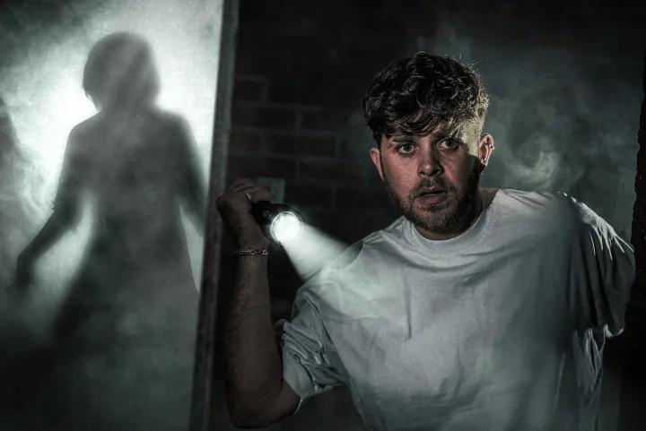 A terrified man holding a flashlight in a smoky, dark room as a shadowy figure looms in the background