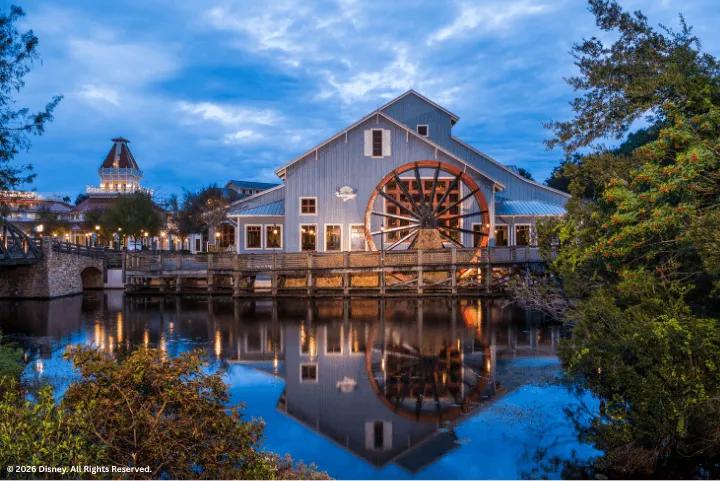 A large wooden water wheel attached to a building reflected in a river at twilight