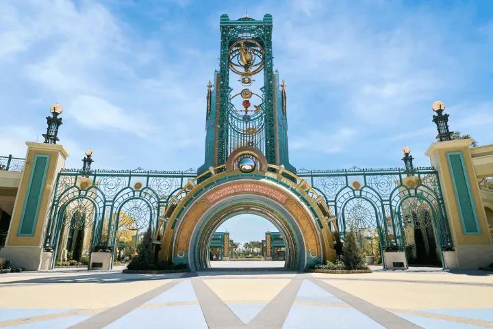 An ornate green and gold metal archway featuring a tall central clock tower under a clear blue sky