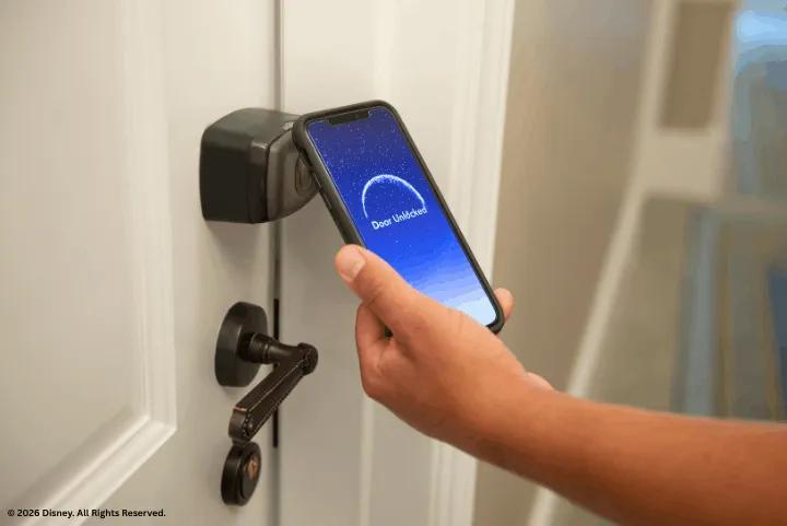 A hand holding a smartphone displaying a Door Unlocked notification against a digital hotel door lock