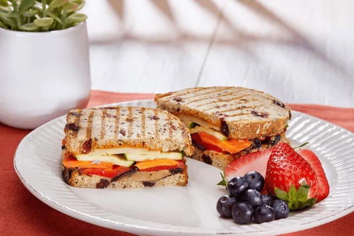 A grilled vegan sandwich cut in half served with fresh strawberries and blueberries on a white plate