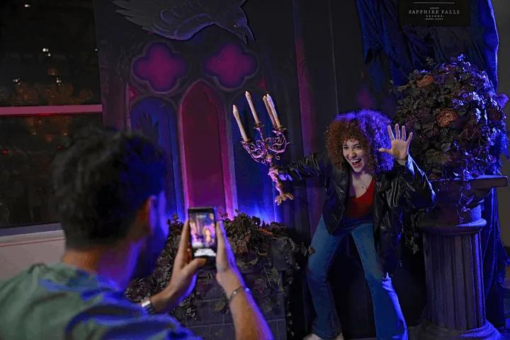 A woman posing in a dark-academia-themed Halloween Horror Nights photo setup at Loews Sapphire Falls Resort while a man takes a picture of her on his smartphone