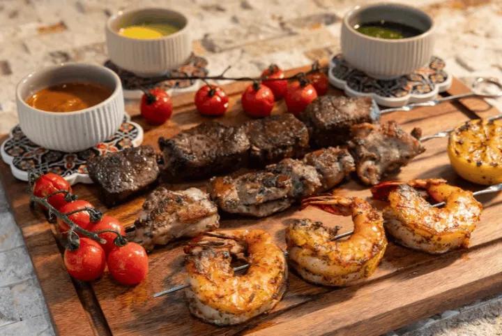 A wooden board topped with skewers of grilled meat and shrimp, alongside tomatoes, grilled lemon, and two dipping sauces