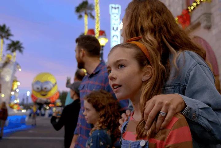 A family stands together on a sidewalk at dusk, watching a giant Minion balloon float by during a holiday parade